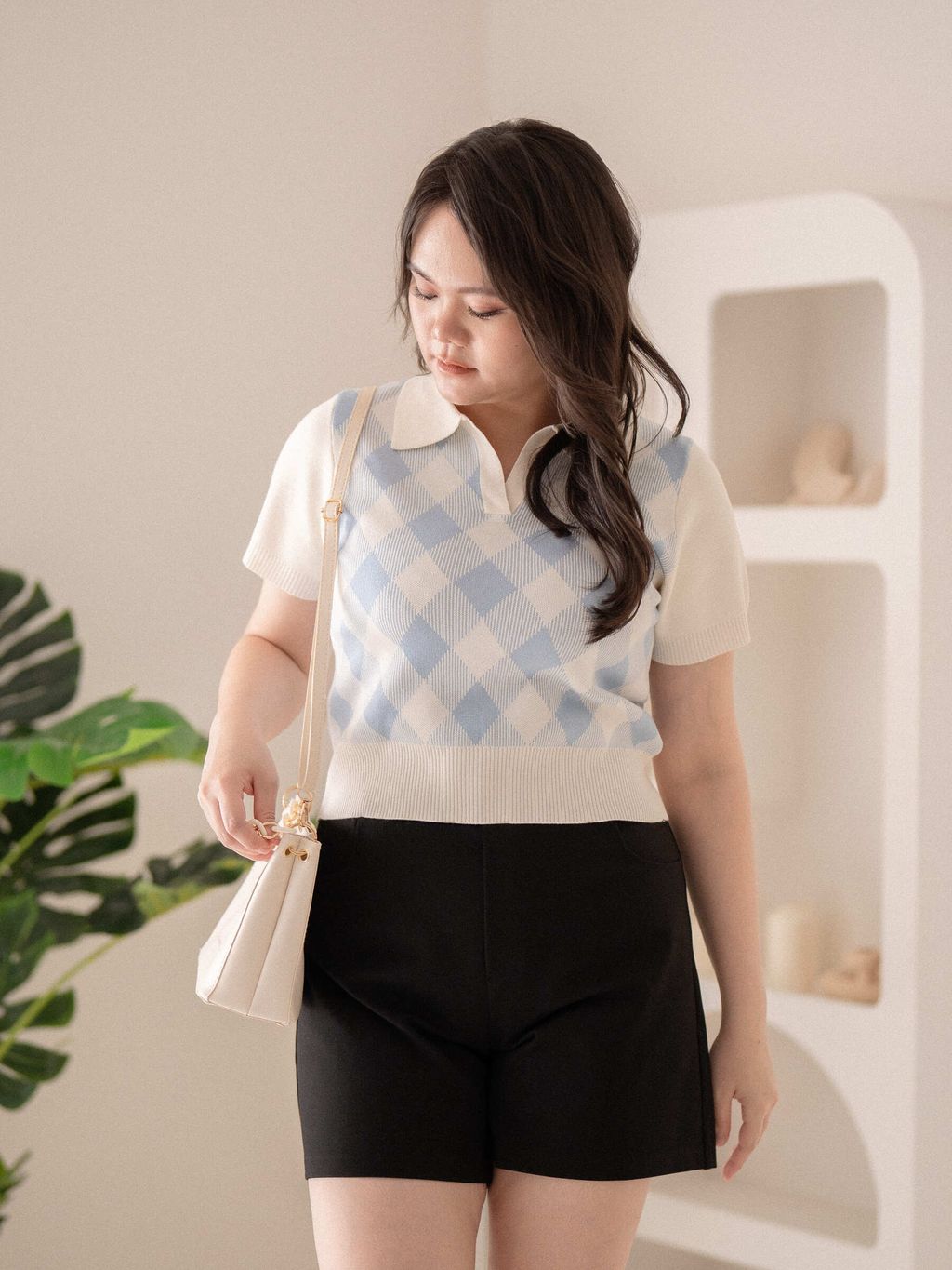 Model wearing blue argyle knit top styled with black shorts and a white bag.