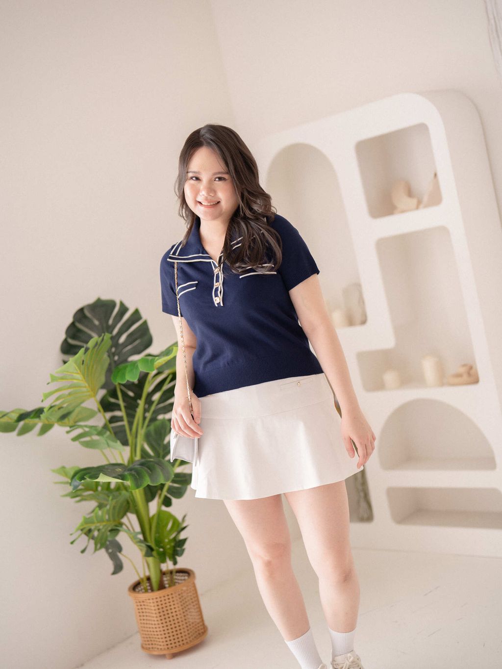 Plus-size model in a dark blue collared knit top with contrast trim and gold buttons, paired with a white flared mini skirt for a fresh and polished summer look.