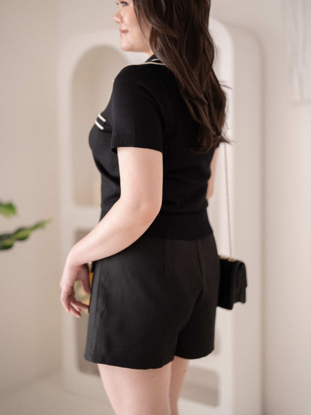 Plus-size woman showcasing the back view of a dark knit polo top with short sleeves and a fitted ribbed waist, paired with black tailored shorts for a sleek silhouette.