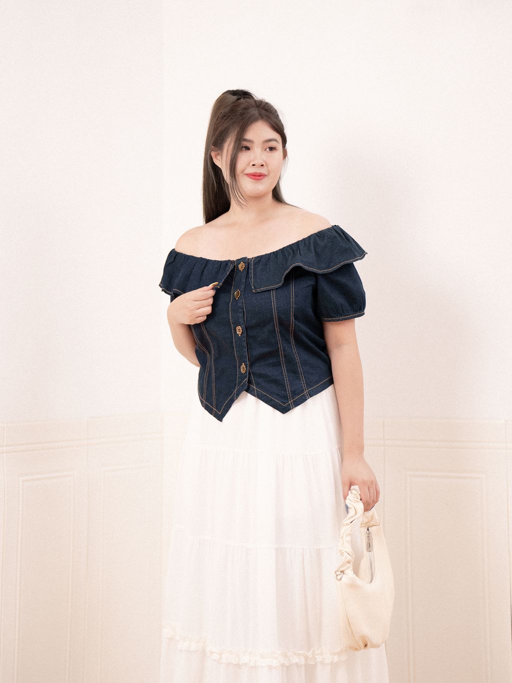 Curvy model showcases the off-shoulder denim blouse with vintage gold buttons and angled hemline, styled with elegance.