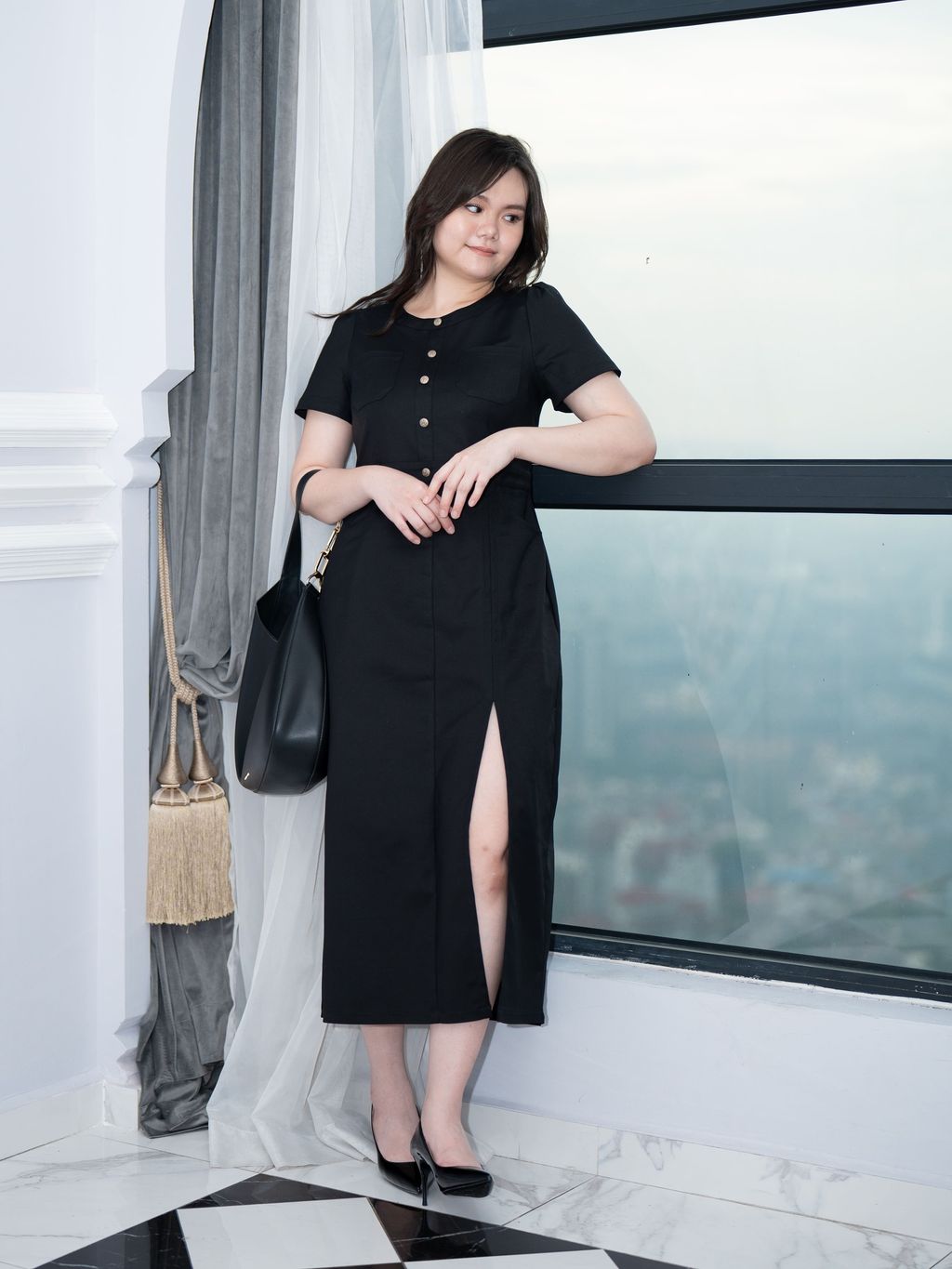 Model in black slit dress standing by window in an elegant indoor setting.