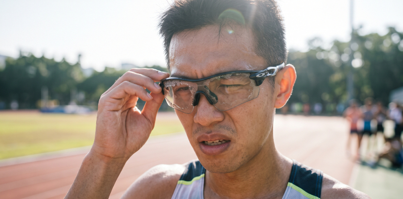 一名運動者因太陽眼鏡鍍膜脫落導致視線模糊而顯露無奈的表情，背景為一片被陽光刺眼的戶外運動場景，突顯運動太陽眼鏡鍍膜脫落的痛點與對視線的影響