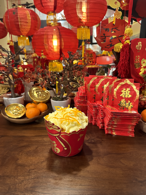 A cupcake with Lunar New Year decorations in the background, including red lanterns and money packets stacked nearby, creating a festive celebration ambiance with warm, inviting lighting.