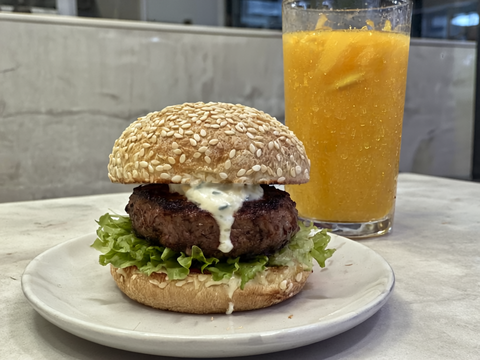 A hamburger with a beef patty, fresh lettuce, and creamy white sauce, served on a plate. Behind the plate, there's a tall glass of cold orange juice, with condensation on the glass, all captured wit - Copy