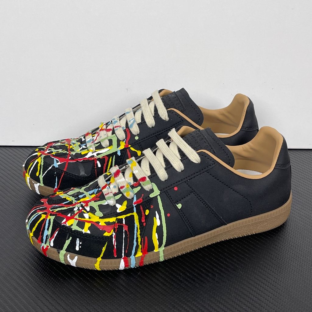 Maison Margiela Replica low-top sneaker in matte black calfskin leather featuring an avant-garde multicolor hand-splattered paint drop design across the toe box and a classic gum sole, style code S58WS0109P1895T1016.