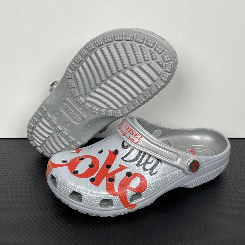 Side profile of the metallic silver Crocs x Diet Coke Classic Clog featuring a large red Coke script graphic on the toe box, a custom Diet Coke hardware rivet, and the retro slogan printed on the pivoting heel strap.