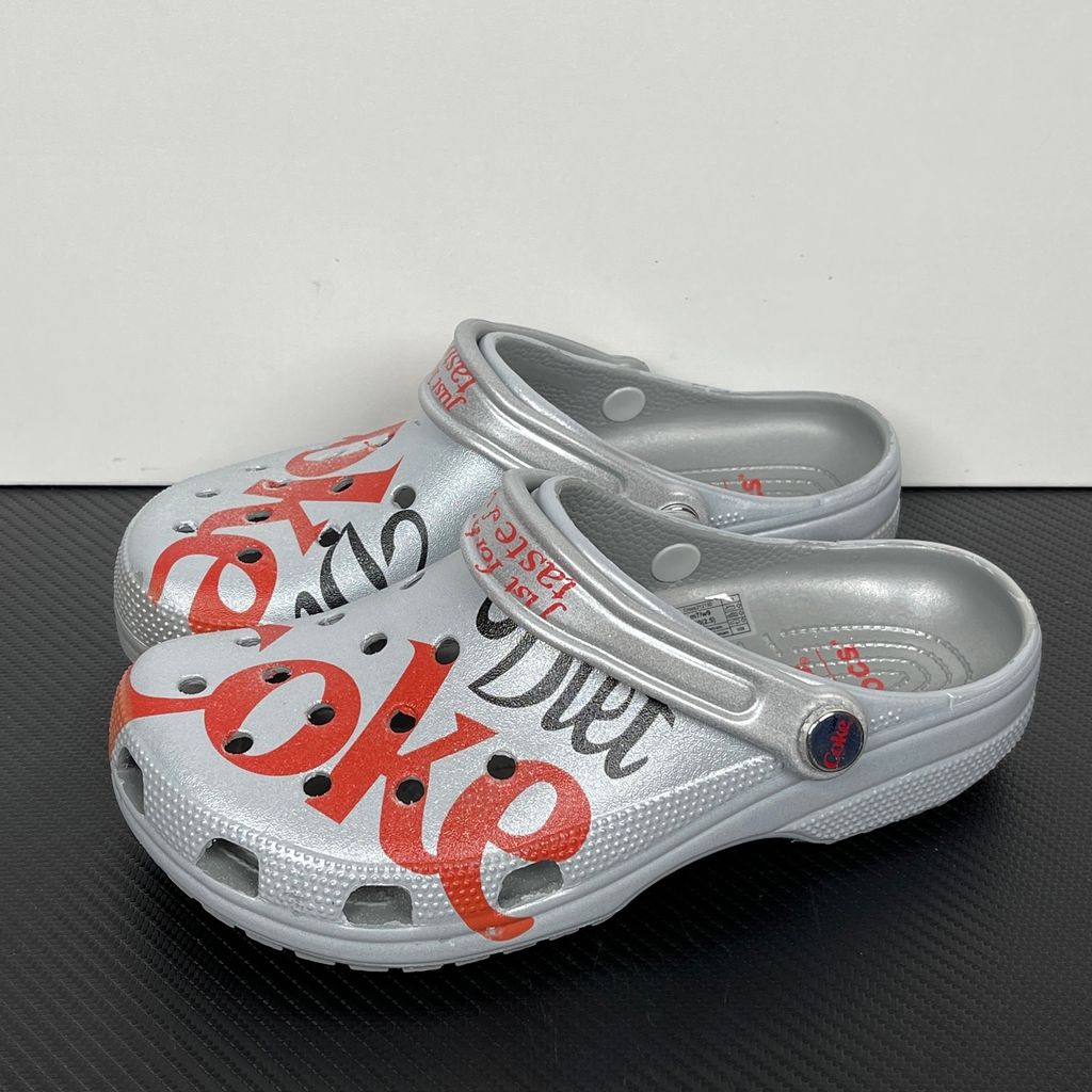 Side profile of the metallic silver Crocs x Diet Coke Classic Clog featuring a large red Coke script graphic on the toe box, a custom Diet Coke hardware rivet, and the retro slogan printed on the pivoting heel strap.