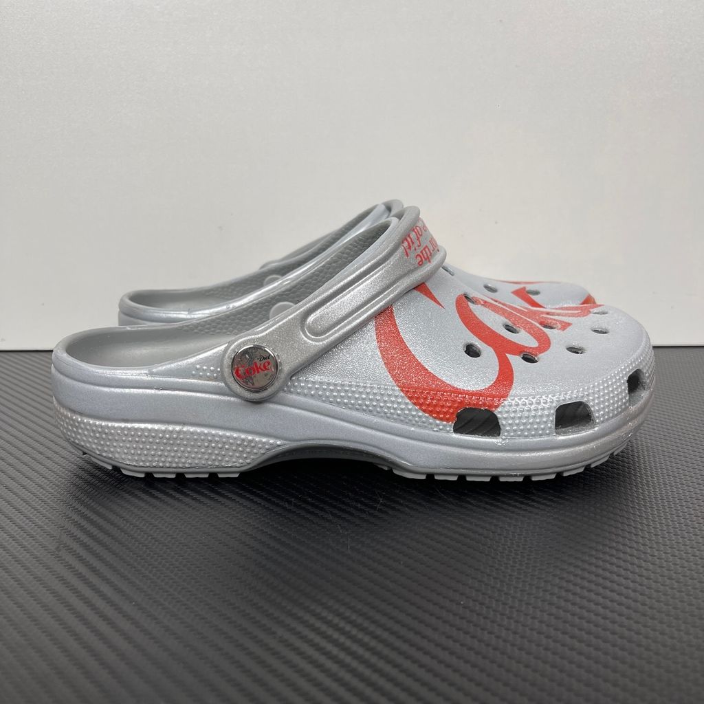 Side profile of the metallic silver Crocs x Diet Coke Classic Clog featuring a large red Coke script graphic on the toe box, a custom Diet Coke hardware rivet, and the retro slogan printed on the pivoting heel strap.