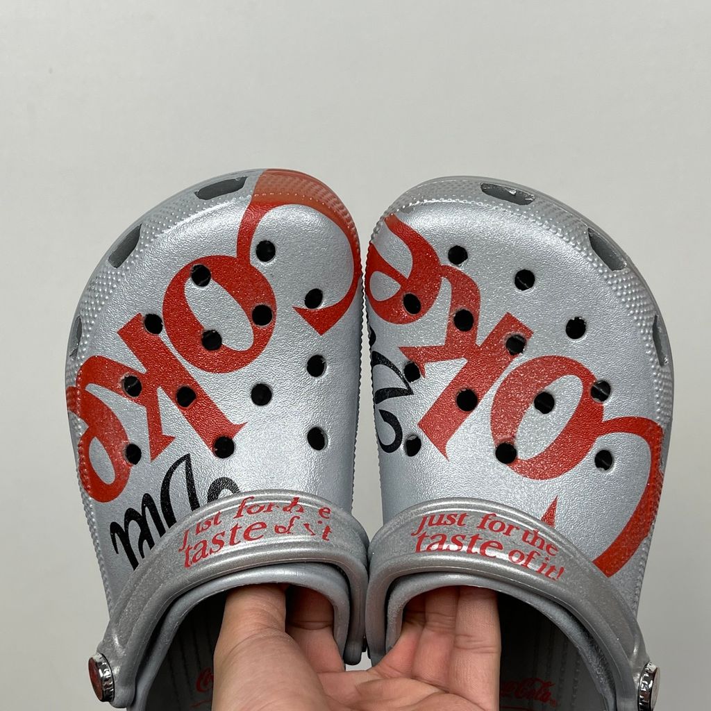 Side profile of the metallic silver Crocs x Diet Coke Classic Clog featuring a large red Coke script graphic on the toe box, a custom Diet Coke hardware rivet, and the retro slogan printed on the pivoting heel strap.