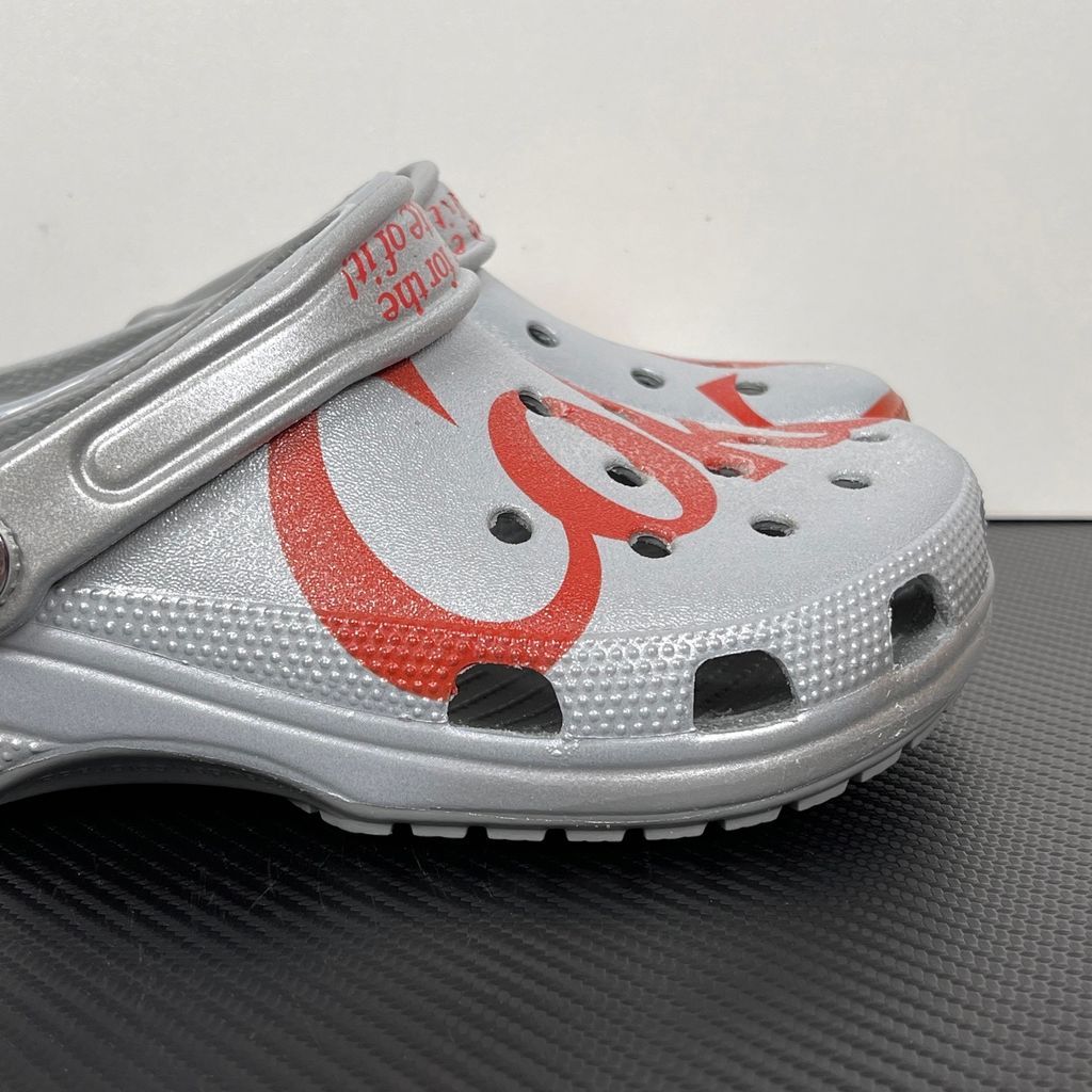 Side profile of the metallic silver Crocs x Diet Coke Classic Clog featuring a large red Coke script graphic on the toe box, a custom Diet Coke hardware rivet, and the retro slogan printed on the pivoting heel strap.