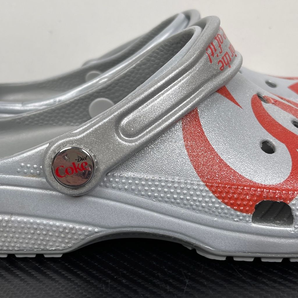 Side profile of the metallic silver Crocs x Diet Coke Classic Clog featuring a large red Coke script graphic on the toe box, a custom Diet Coke hardware rivet, and the retro slogan printed on the pivoting heel strap.