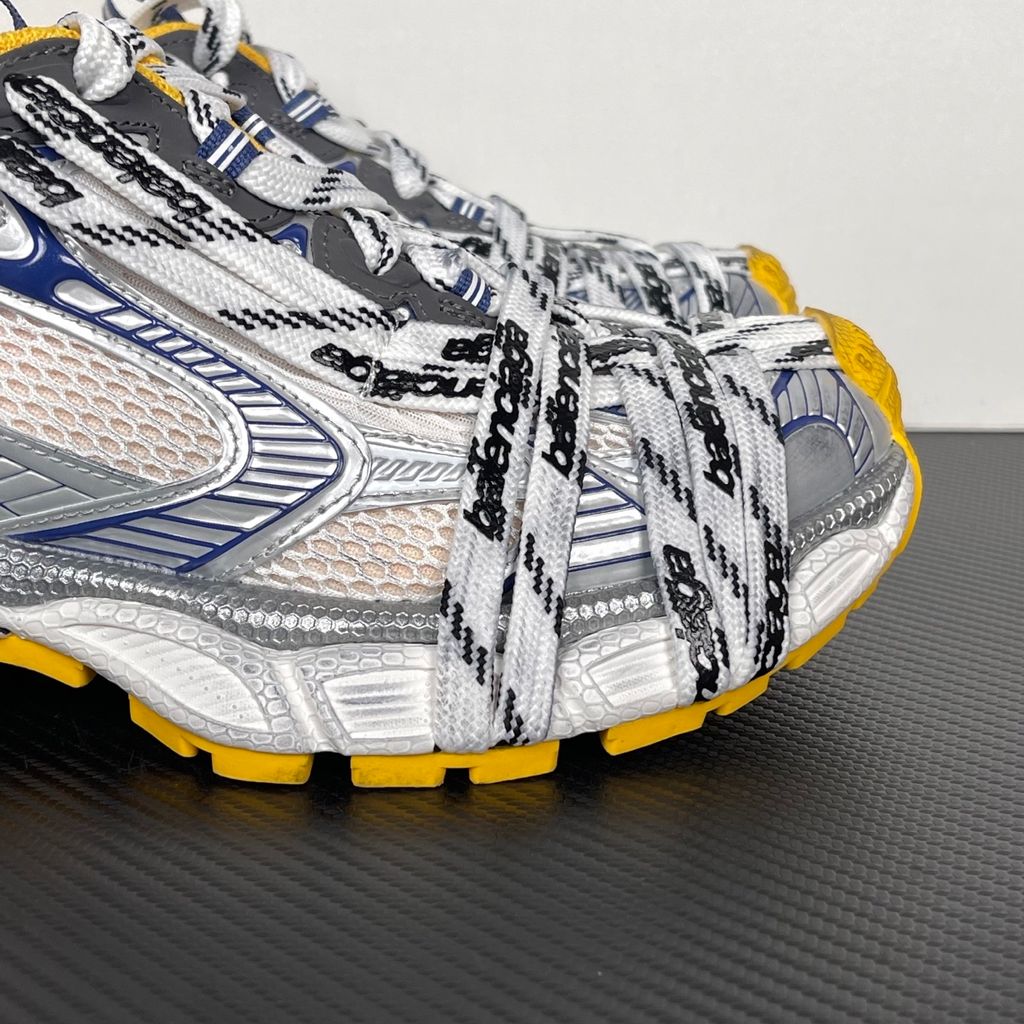 Balenciaga 3XL Extreme Laces sneaker side profile, featuring exaggerated logo-printed laces wrapped around a silver, blue, and white mesh upper with a vibrant yellow-orange outsole.