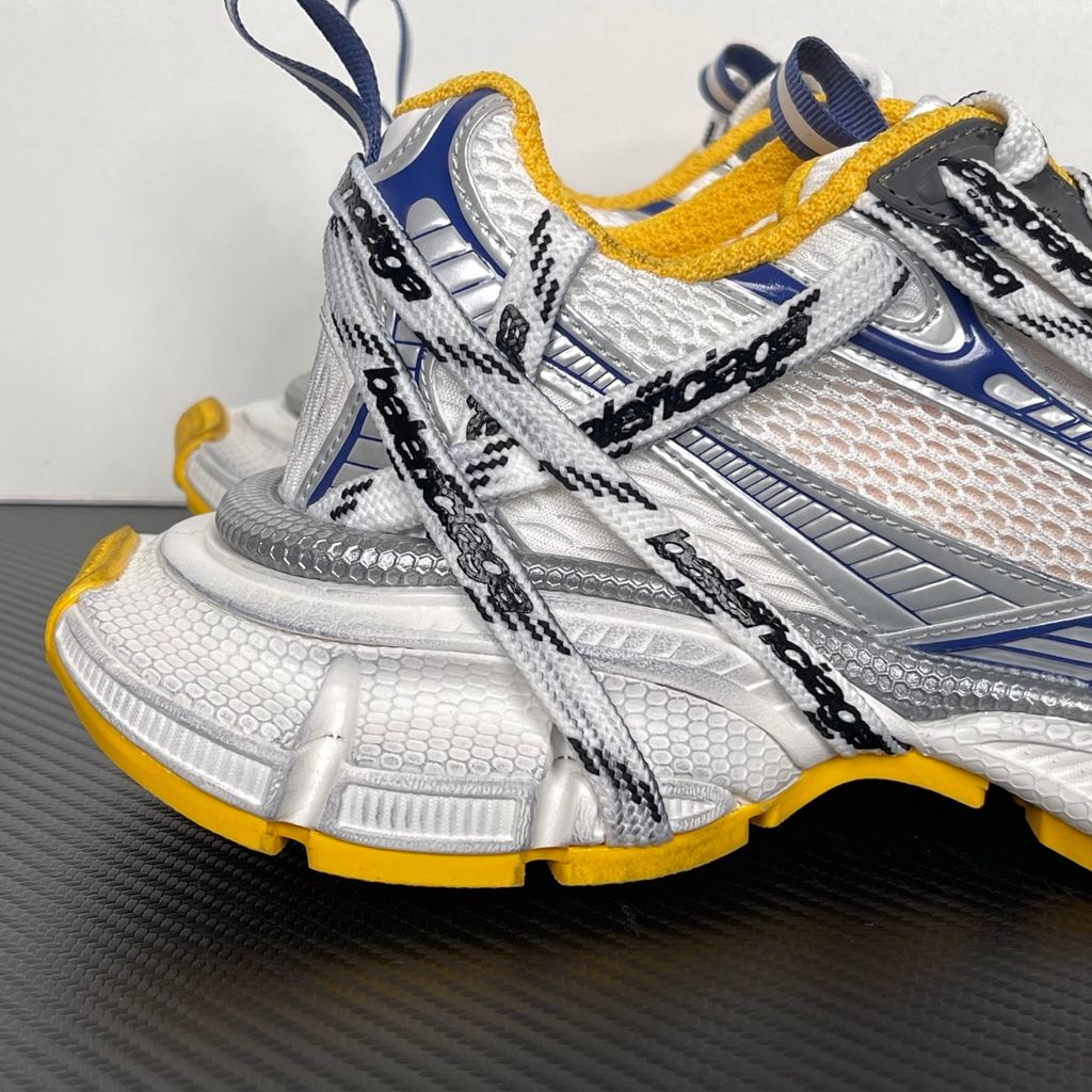 Balenciaga 3XL Extreme Laces sneaker side profile, featuring exaggerated logo-printed laces wrapped around a silver, blue, and white mesh upper with a vibrant yellow-orange outsole.