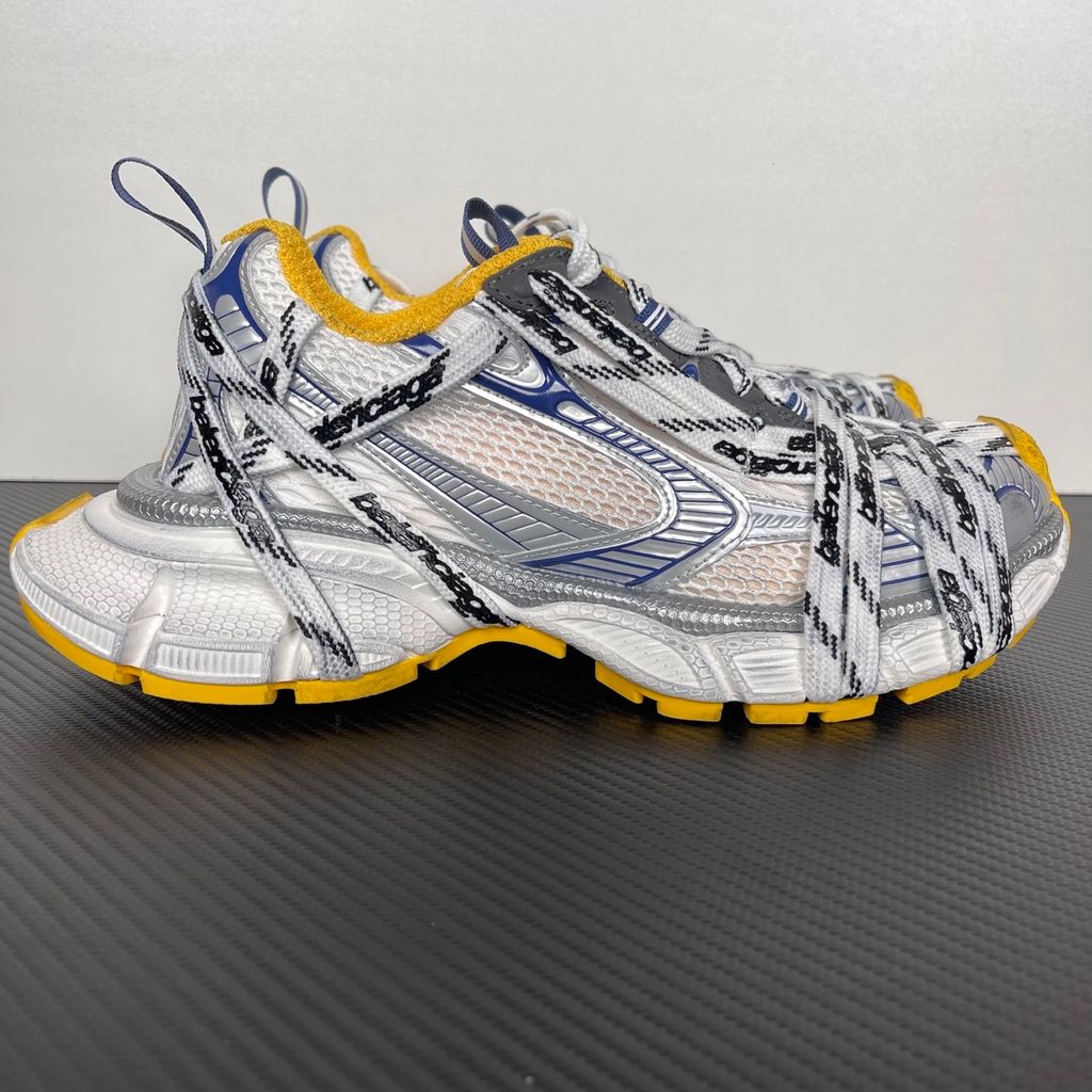 Balenciaga 3XL Extreme Laces sneaker side profile, featuring exaggerated logo-printed laces wrapped around a silver, blue, and white mesh upper with a vibrant yellow-orange outsole.