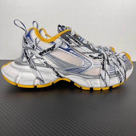 Balenciaga 3XL Extreme Laces sneaker side profile, featuring exaggerated logo-printed laces wrapped around a silver, blue, and white mesh upper with a vibrant yellow-orange outsole.