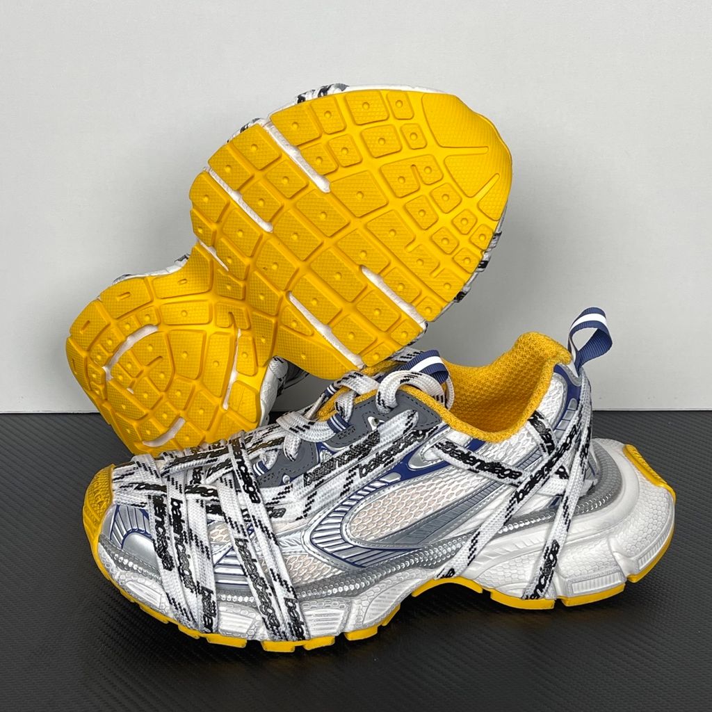 Balenciaga 3XL Extreme Laces sneaker side profile, featuring exaggerated logo-printed laces wrapped around a silver, blue, and white mesh upper with a vibrant yellow-orange outsole.