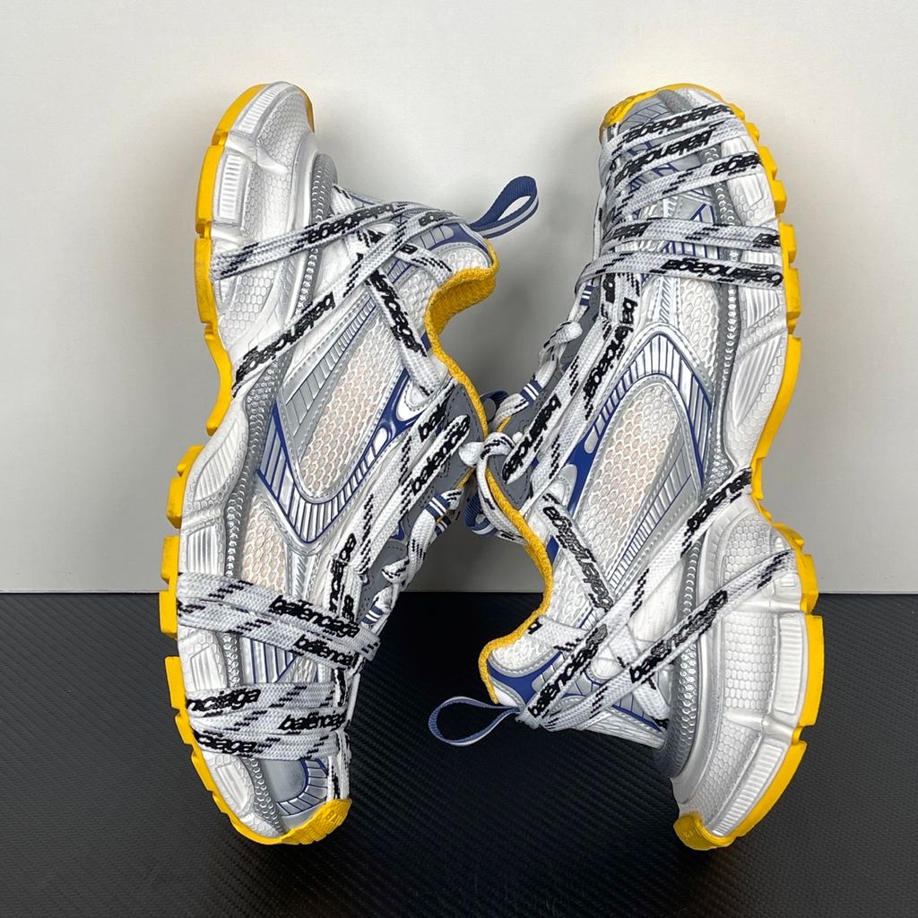 Balenciaga 3XL Extreme Laces sneaker side profile, featuring exaggerated logo-printed laces wrapped around a silver, blue, and white mesh upper with a vibrant yellow-orange outsole.