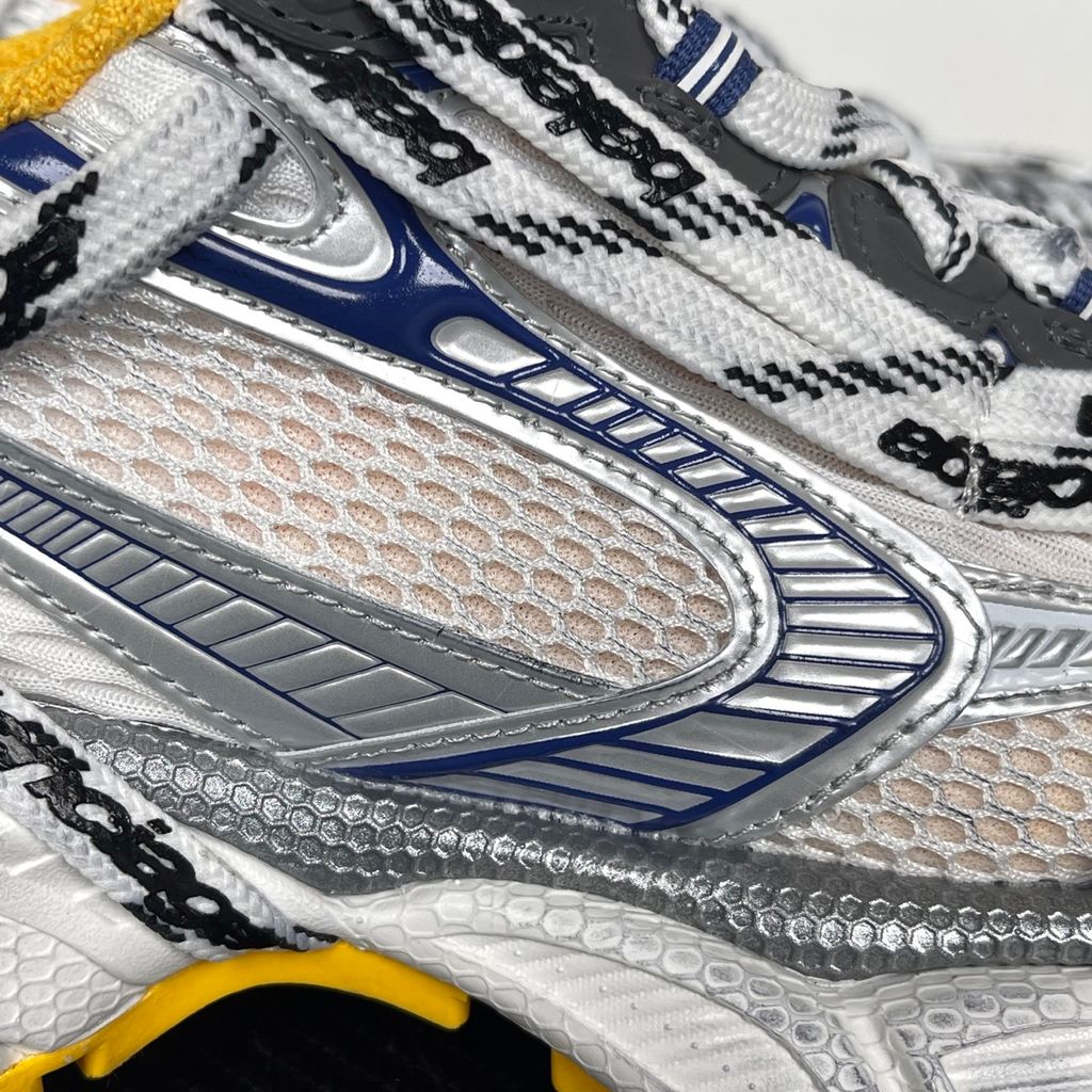 Balenciaga 3XL Extreme Laces sneaker side profile, featuring exaggerated logo-printed laces wrapped around a silver, blue, and white mesh upper with a vibrant yellow-orange outsole.