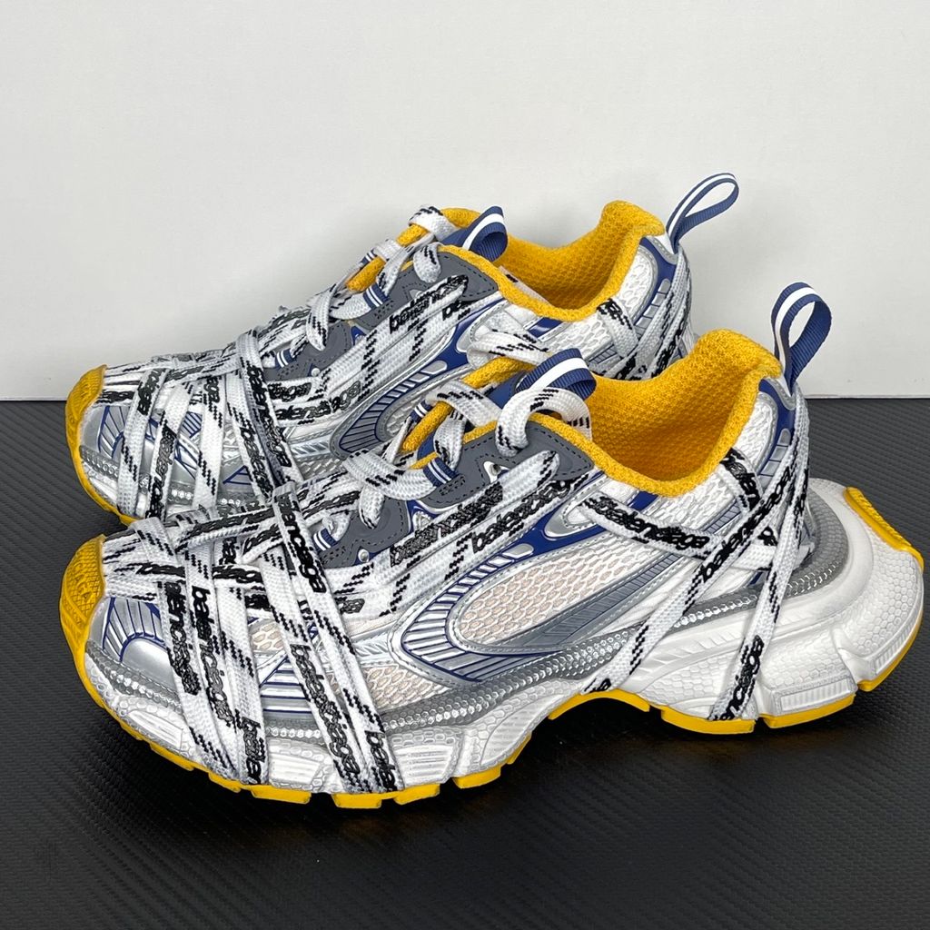 Balenciaga 3XL Extreme Laces sneaker side profile, featuring exaggerated logo-printed laces wrapped around a silver, blue, and white mesh upper with a vibrant yellow-orange outsole.