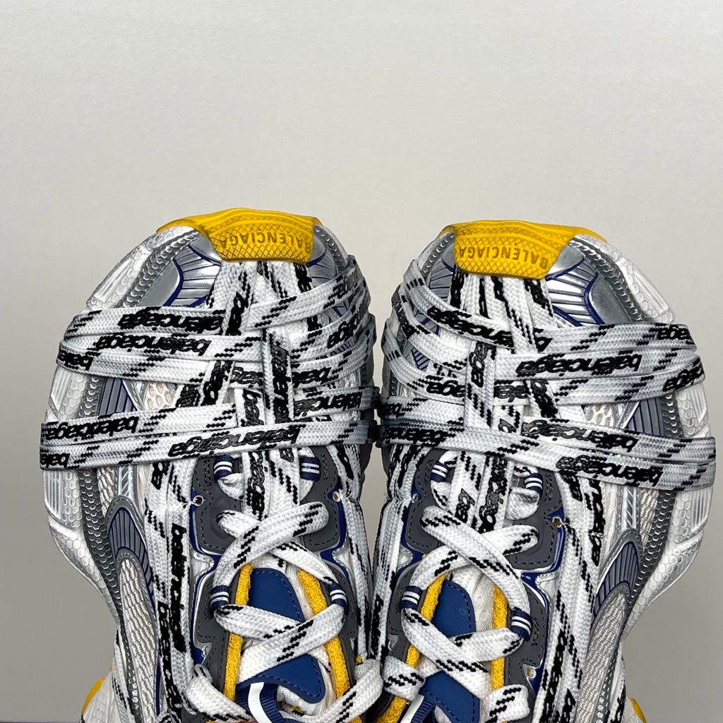 Balenciaga 3XL Extreme Laces sneaker side profile, featuring exaggerated logo-printed laces wrapped around a silver, blue, and white mesh upper with a vibrant yellow-orange outsole.