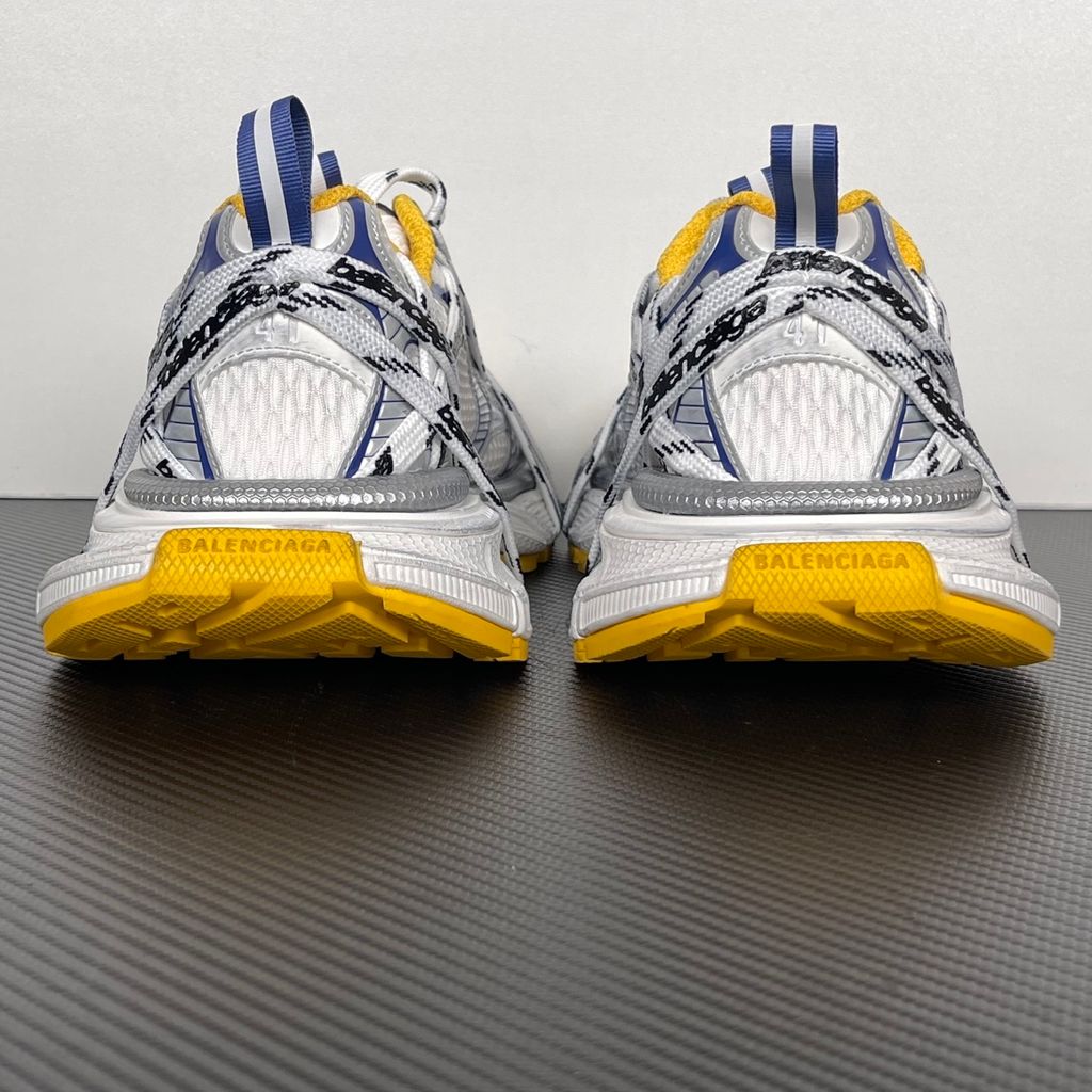 Balenciaga 3XL Extreme Laces sneaker side profile, featuring exaggerated logo-printed laces wrapped around a silver, blue, and white mesh upper with a vibrant yellow-orange outsole.