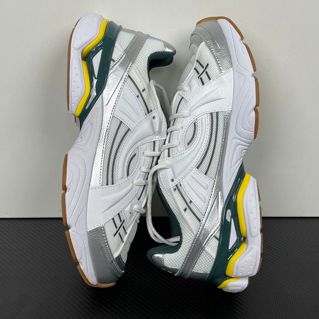 Under Armour UA Sola retro tech-runner side profile, featuring a clean white breathable mesh upper, dynamic metallic silver overlays, dark green collar lining, bright yellow midsole accents, and a classic gum rubber outsole, SKU 6005284-104.
