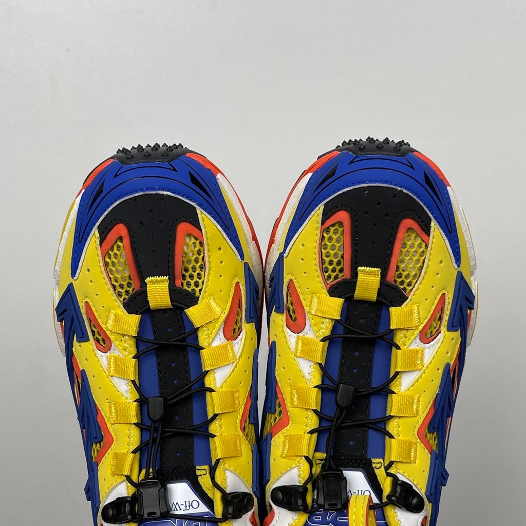 Side profile of the Off-White "Be Right" sneaker featuring a vibrant yellow mesh upper, bold blue signature arrow overlays, a chunky orange sculpted midsole, and black technical heel straps.