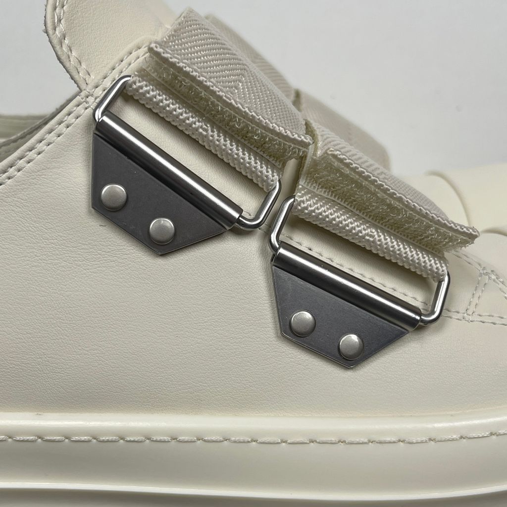 Side profile of the Rick Owens Mainline low-top sneaker in off-white leather, featuring distinctive industrial metal hardware, textured velcro strap closures, and a chunky tonal rubber sole.