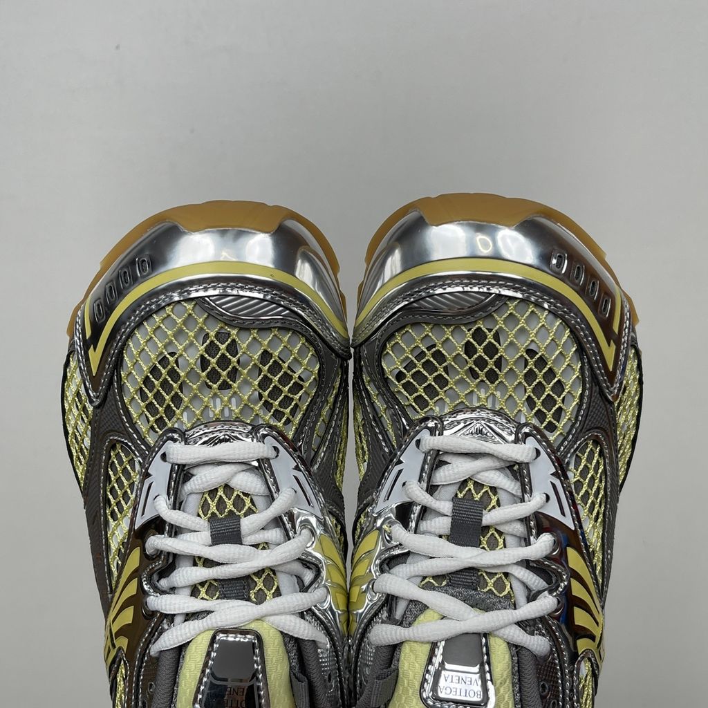 Side profile of the Bottega Veneta Orbit Runner sneaker, showcasing its intricate metallic and green-yellow technical mesh construction, prominent "BOTTEGA VENETA" branding, and the textured gum-tone outsole.