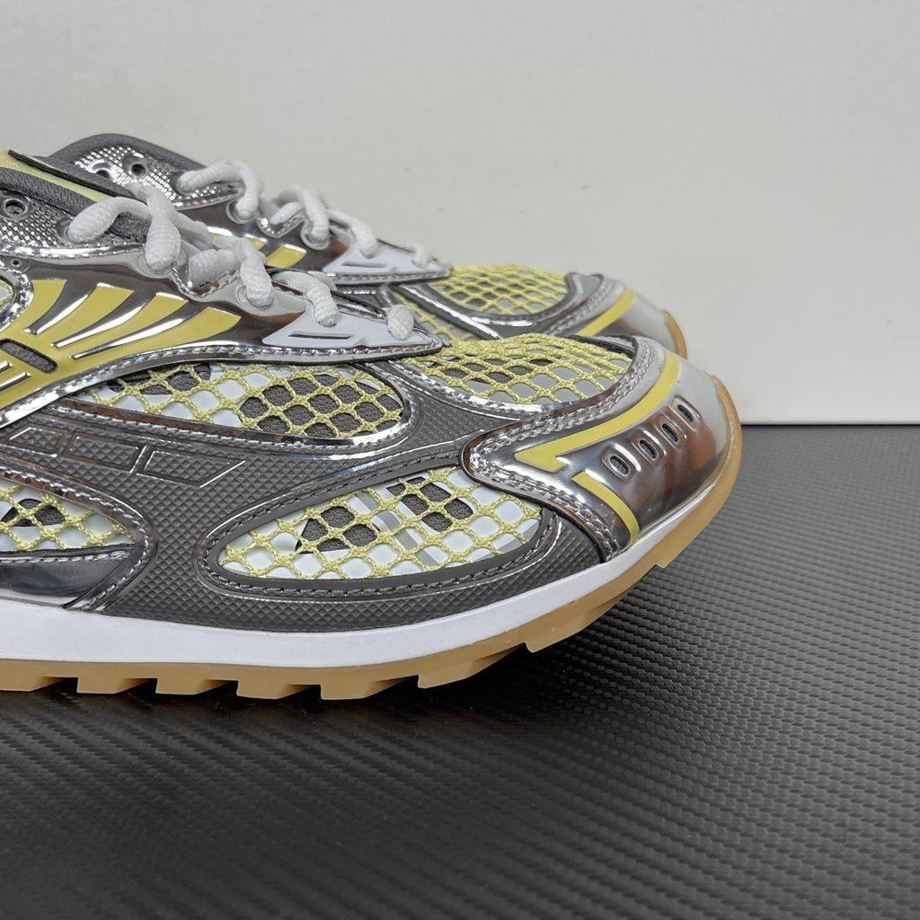 Side profile of the Bottega Veneta Orbit Runner sneaker, showcasing its intricate metallic and green-yellow technical mesh construction, prominent "BOTTEGA VENETA" branding, and the textured gum-tone outsole.