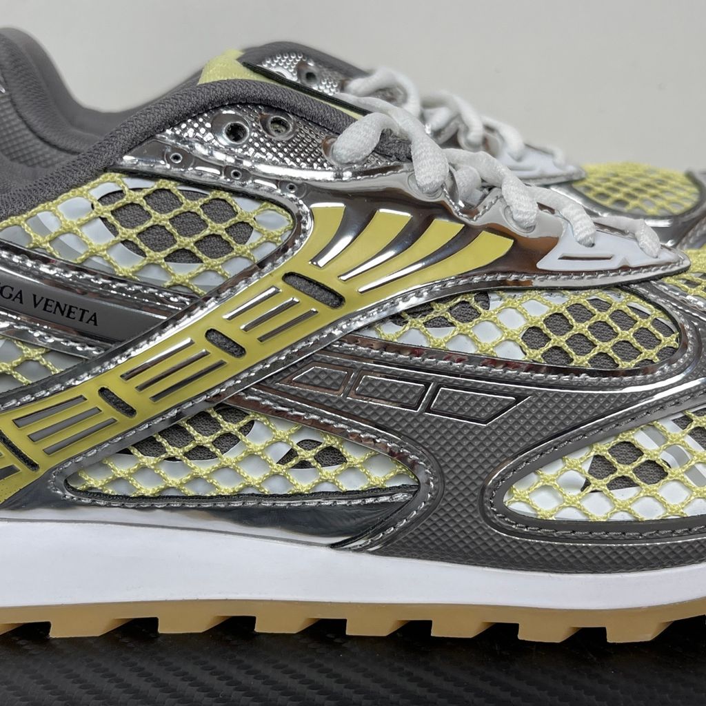 Side profile of the Bottega Veneta Orbit Runner sneaker, showcasing its intricate metallic and green-yellow technical mesh construction, prominent "BOTTEGA VENETA" branding, and the textured gum-tone outsole.