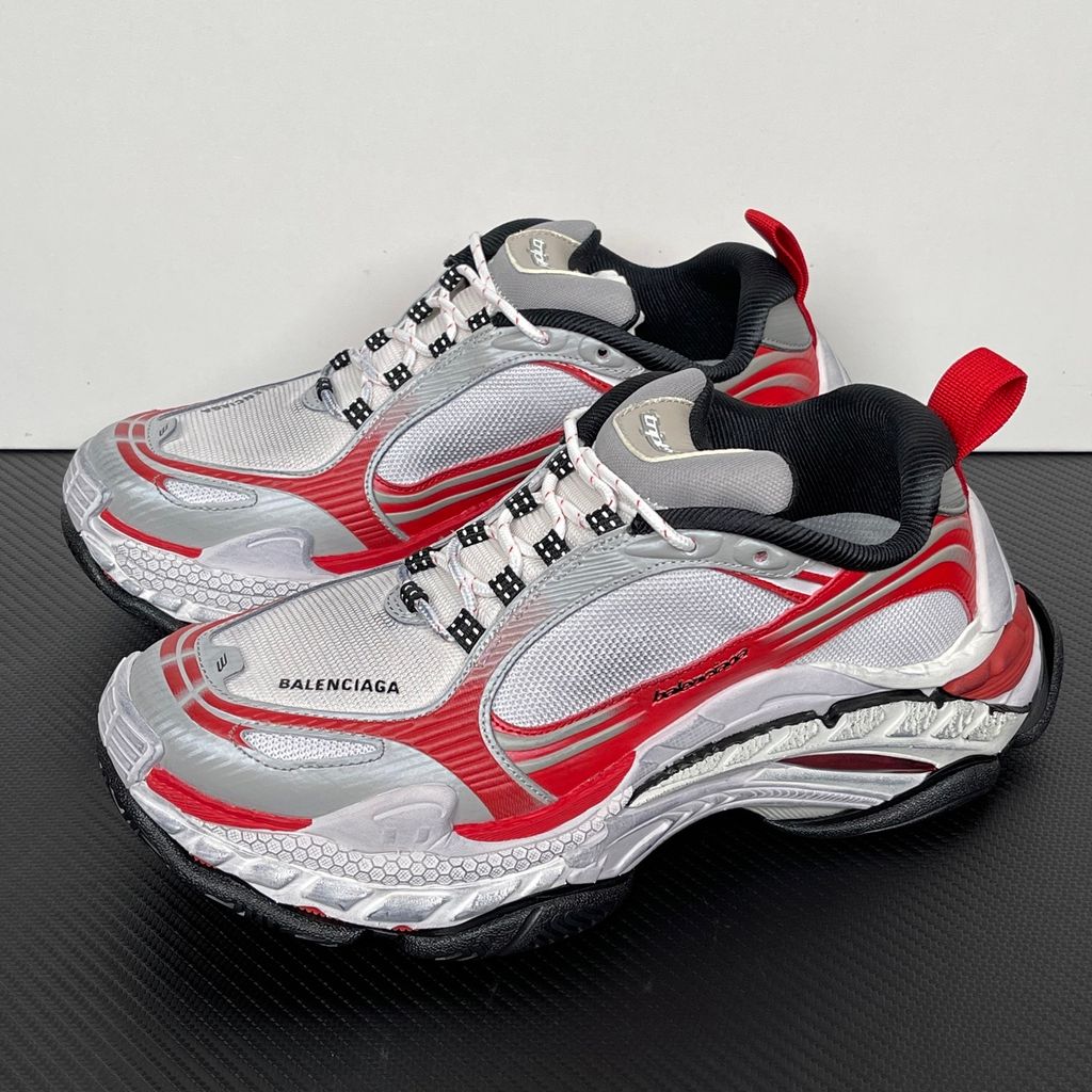 Side profile of the Balenciaga Triple S 2 sneaker, showcasing the complex layered mesh and synthetic upper in grey, red, and white, alongside the signature distressed chunky, multi-layered rubber sole.