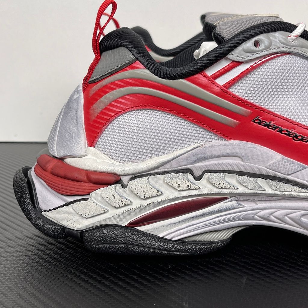 Side profile of the Balenciaga Triple S 2 sneaker, showcasing the complex layered mesh and synthetic upper in grey, red, and white, alongside the signature distressed chunky, multi-layered rubber sole.