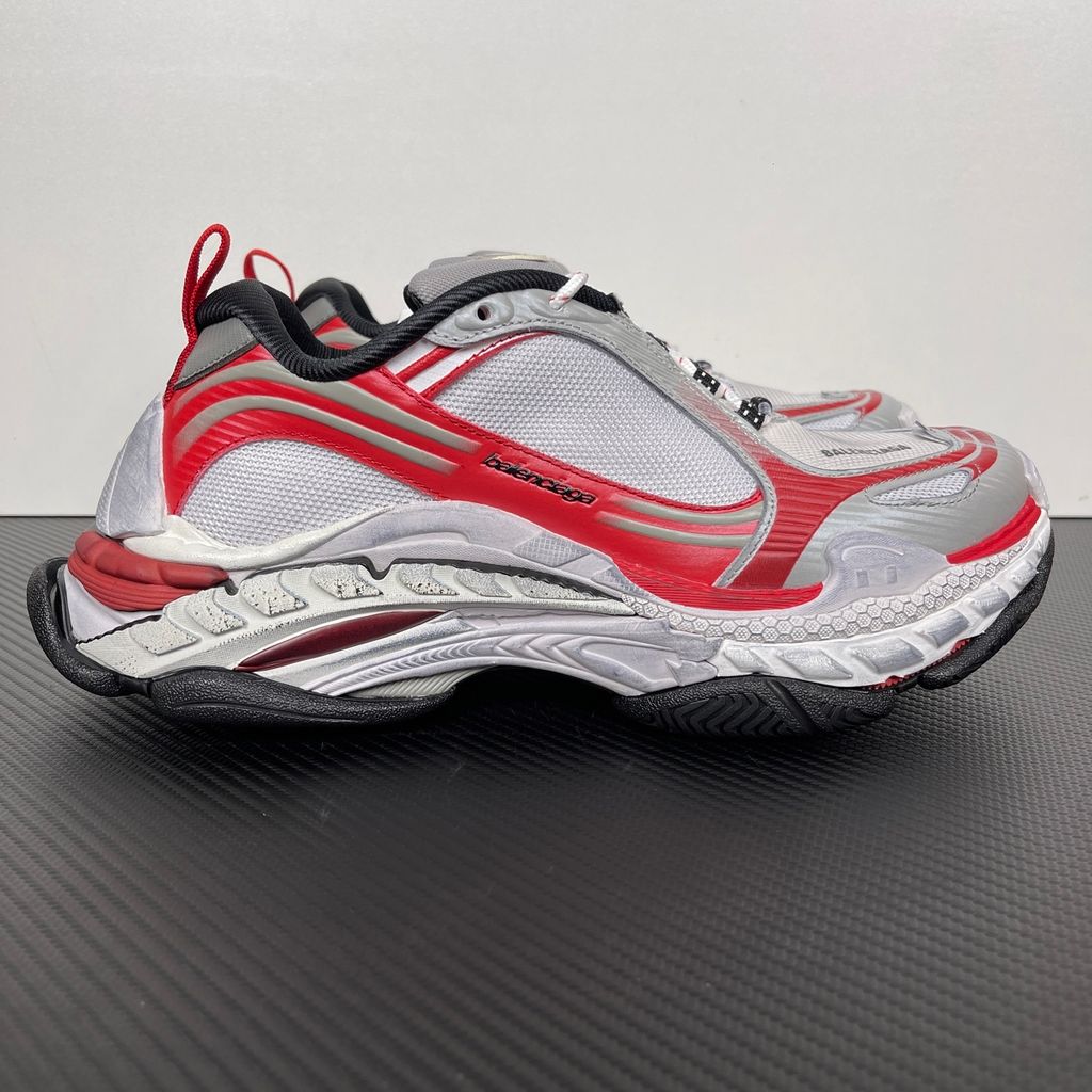Side profile of the Balenciaga Triple S 2 sneaker, showcasing the complex layered mesh and synthetic upper in grey, red, and white, alongside the signature distressed chunky, multi-layered rubber sole.