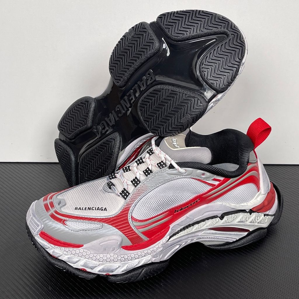 Side profile of the Balenciaga Triple S 2 sneaker, showcasing the complex layered mesh and synthetic upper in grey, red, and white, alongside the signature distressed chunky, multi-layered rubber sole.
