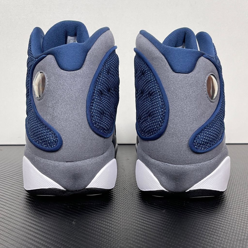 Side profile of the Air Jordan 13 Retro 'Flint' (2026), showcasing the reflective navy blue dimpled mesh upper, Flint Grey suede mudguard, clean white leather toe, and the iconic green holographic cat eye emblem on the heel.