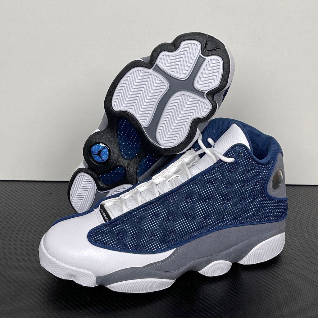 Side profile of the Air Jordan 13 Retro 'Flint' (2026), showcasing the reflective navy blue dimpled mesh upper, Flint Grey suede mudguard, clean white leather toe, and the iconic green holographic cat eye emblem on the heel.