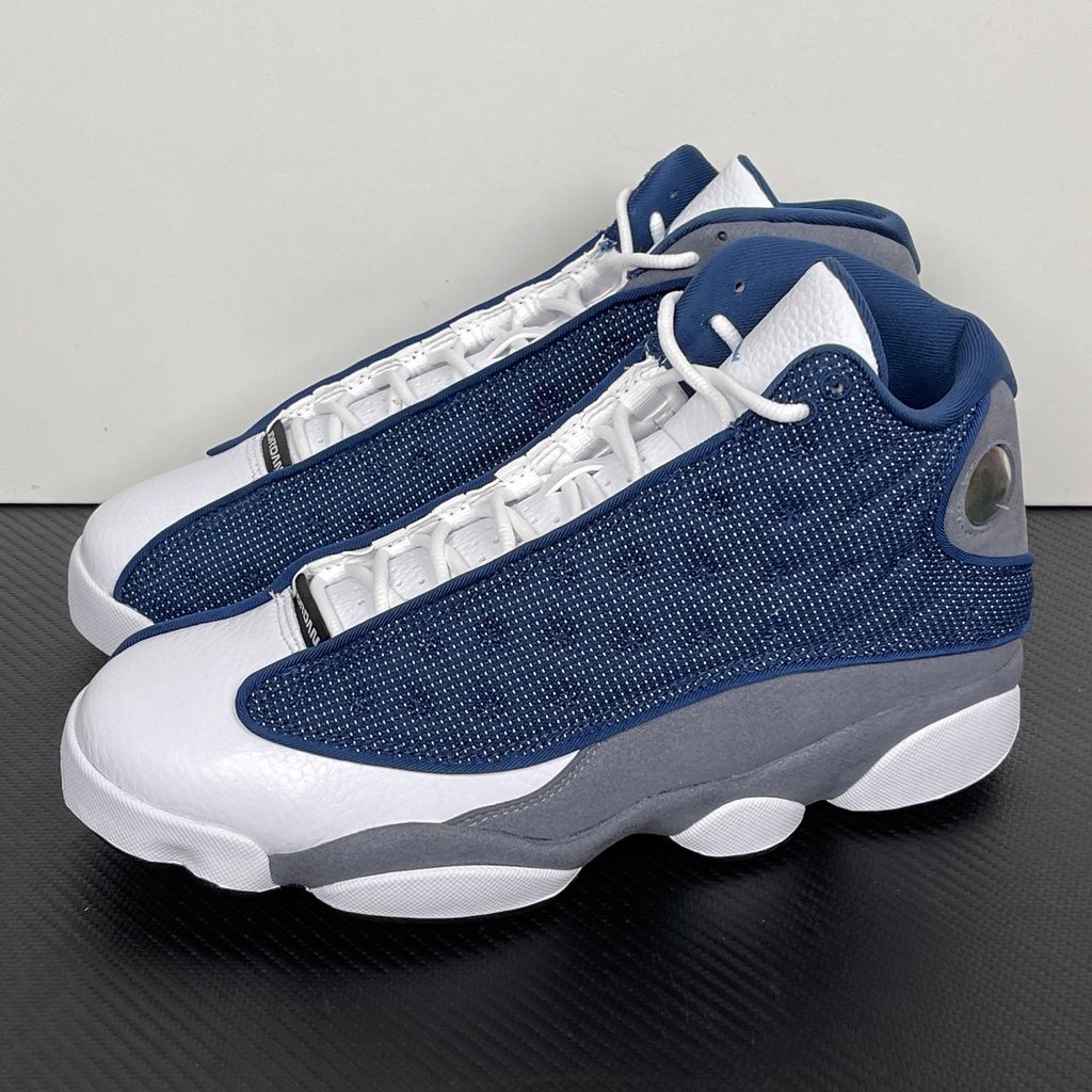 Side profile of the Air Jordan 13 Retro 'Flint' (2026), showcasing the reflective navy blue dimpled mesh upper, Flint Grey suede mudguard, clean white leather toe, and the iconic green holographic cat eye emblem on the heel.