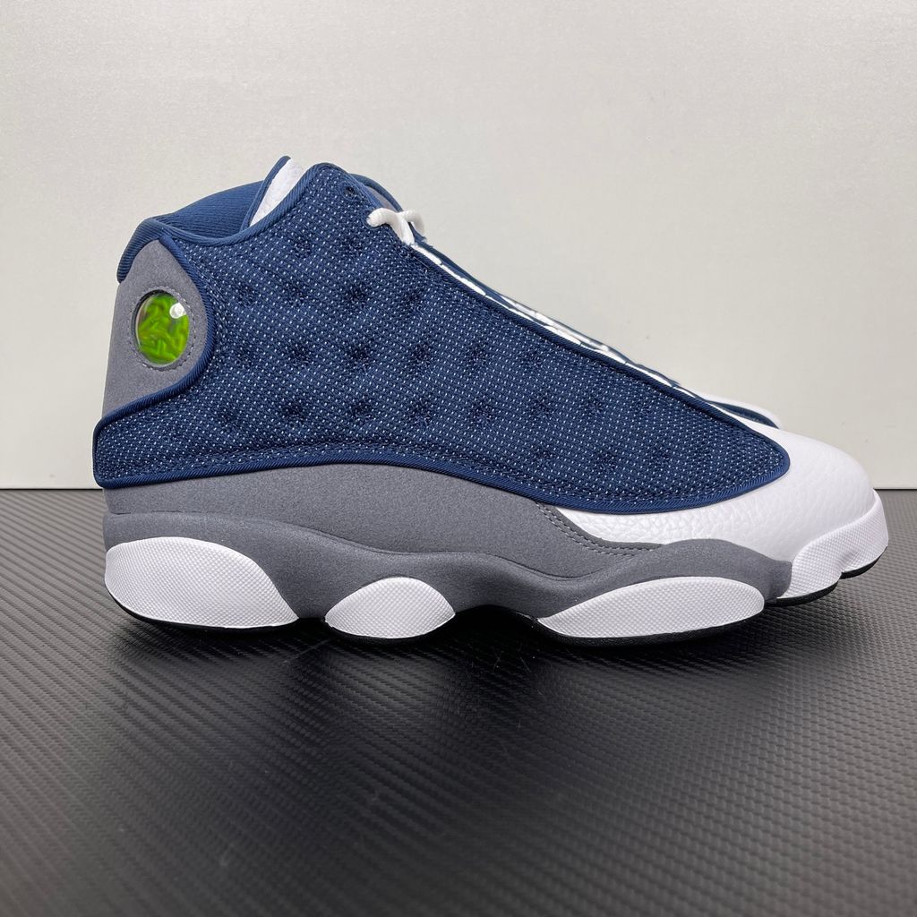 Side profile of the Air Jordan 13 Retro 'Flint' (2026), showcasing the reflective navy blue dimpled mesh upper, Flint Grey suede mudguard, clean white leather toe, and the iconic green holographic cat eye emblem on the heel.