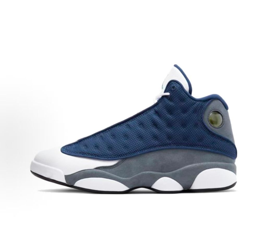 Side profile of the Air Jordan 13 Retro 'Flint' (2026), showcasing the reflective navy blue dimpled mesh upper, Flint Grey suede mudguard, clean white leather toe, and the iconic green holographic cat eye emblem on the heel.