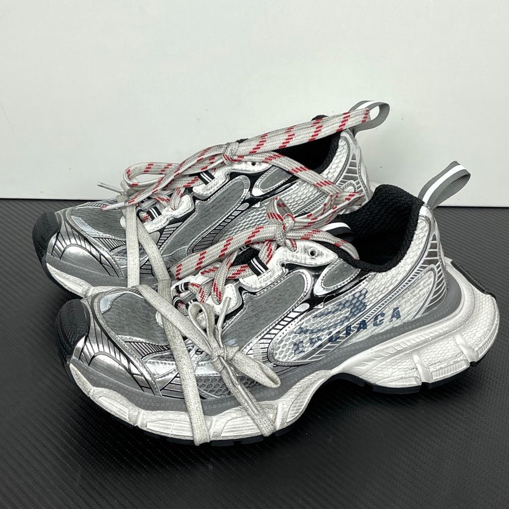 Side profile of the Balenciaga 3XL Sneakers in a distressed silver and white colorway, showcasing the complex metallic overlays, wide mesh base, exaggerated chunky runner sole, and the signature extra laces wrapped around the toe box.