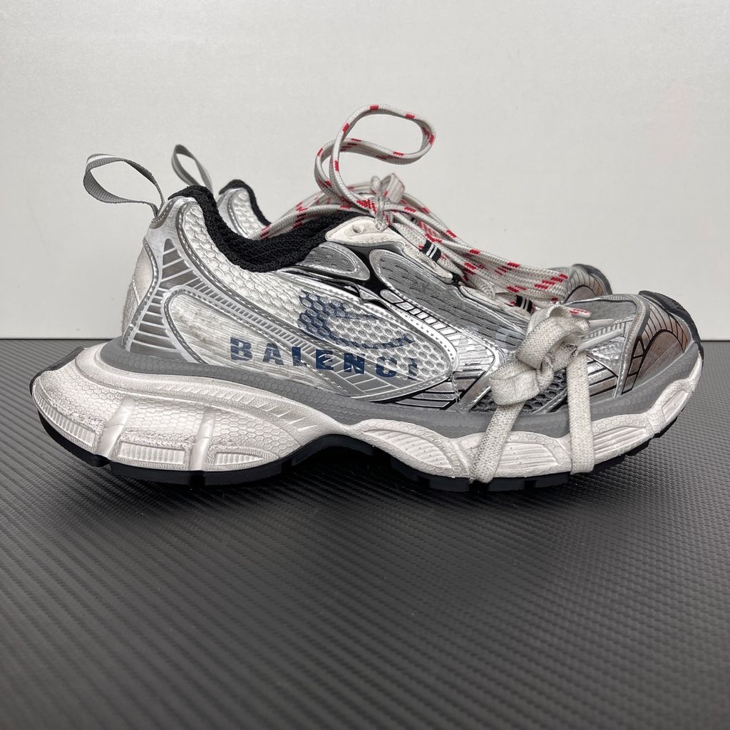 Side profile of the Balenciaga 3XL Sneakers in a distressed silver and white colorway, showcasing the complex metallic overlays, wide mesh base, exaggerated chunky runner sole, and the signature extra laces wrapped around the toe box.