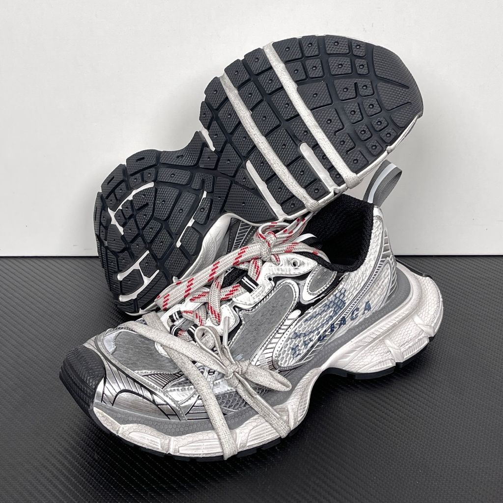 Side profile of the Balenciaga 3XL Sneakers in a distressed silver and white colorway, showcasing the complex metallic overlays, wide mesh base, exaggerated chunky runner sole, and the signature extra laces wrapped around the toe box.