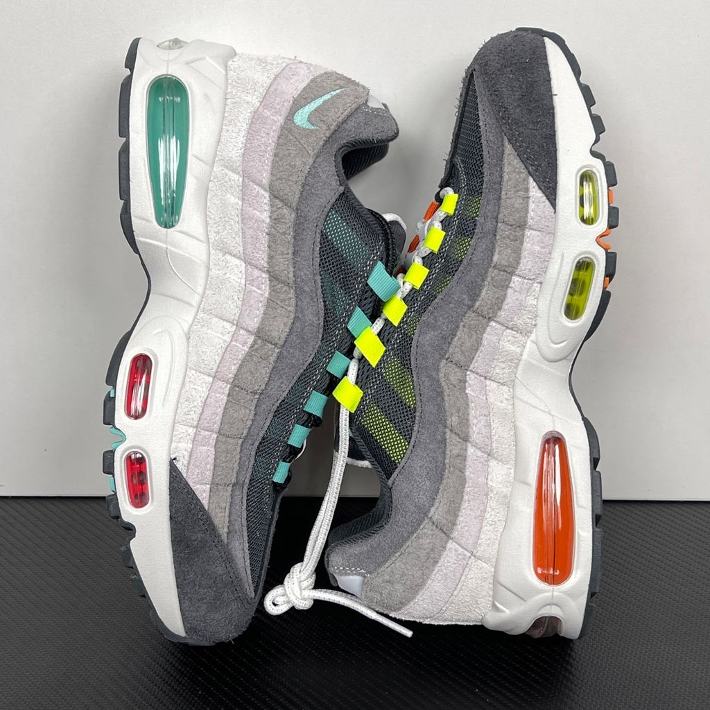 Side profiles of the left and right Nike Air Max 95 OG 'Big Bubble' Greedy (2026) sneakers, showcasing the asymmetrical, mismatched multi-color gradient uppers and the exaggerated visible Air units in the black midsole.