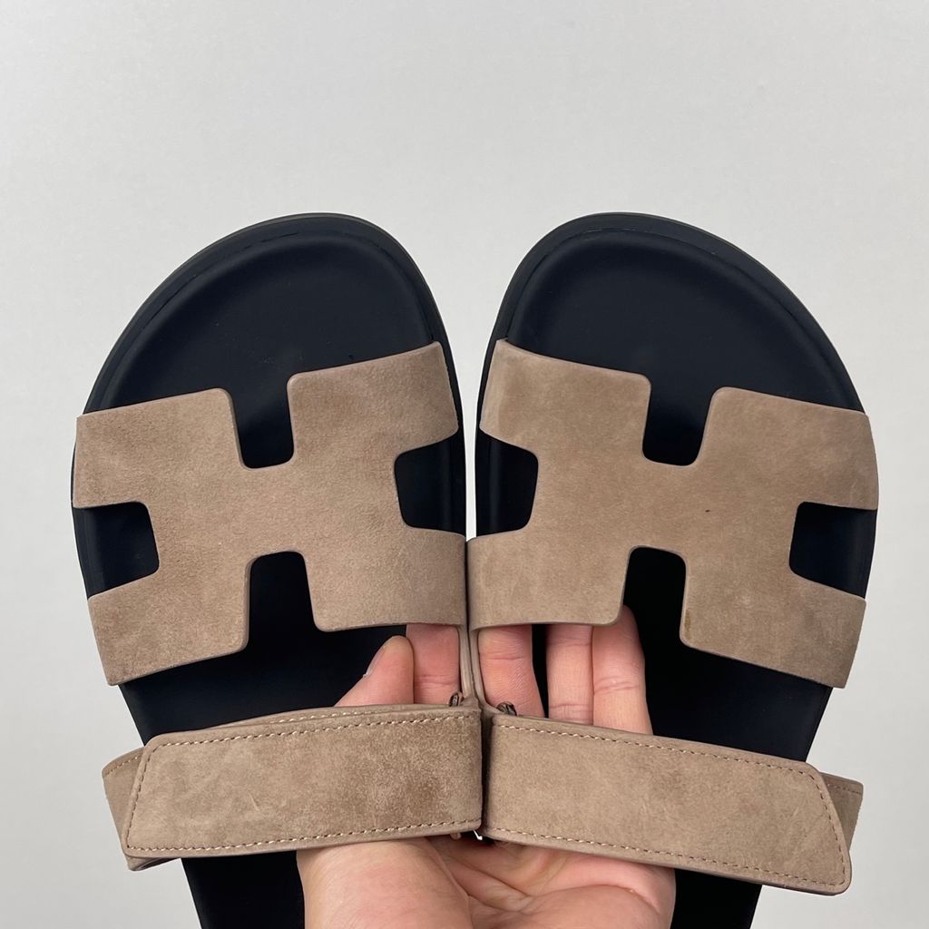Angled top-down view of the Hermès Genius Sandals in taupe grey suede, showcasing the signature 'H' cutout front strap, the adjustable hook-and-loop instep strap, and the contoured black ergonomic rubber footbed with Hermès branding.