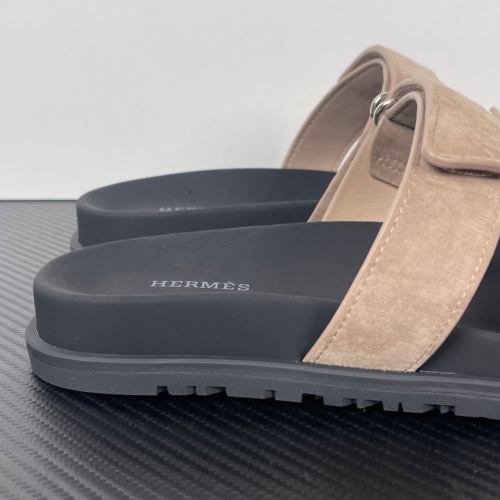 Angled top-down view of the Hermès Genius Sandals in taupe grey suede, showcasing the signature 'H' cutout front strap, the adjustable hook-and-loop instep strap, and the contoured black ergonomic rubber footbed with Hermès branding.