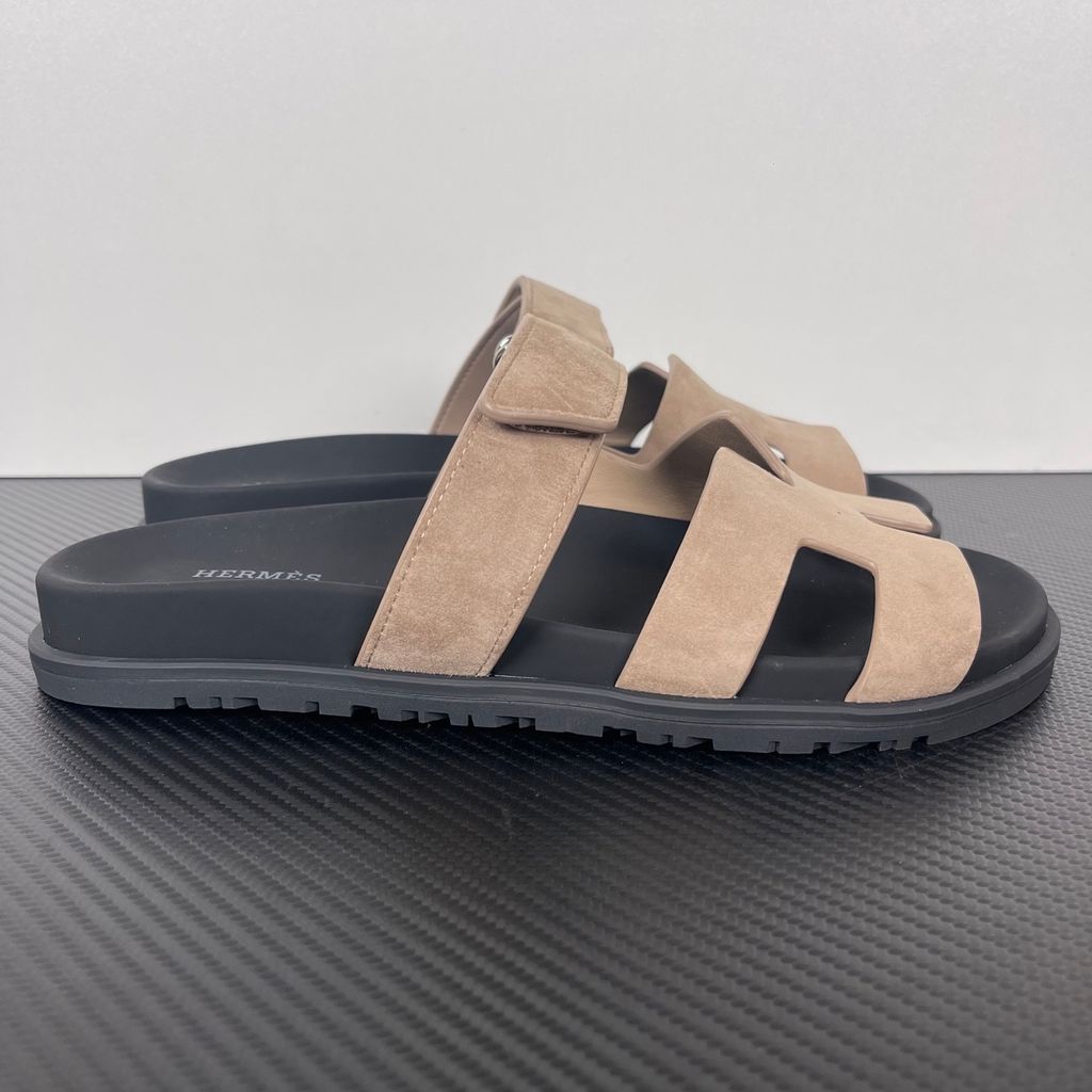 Angled top-down view of the Hermès Genius Sandals in taupe grey suede, showcasing the signature 'H' cutout front strap, the adjustable hook-and-loop instep strap, and the contoured black ergonomic rubber footbed with Hermès branding.