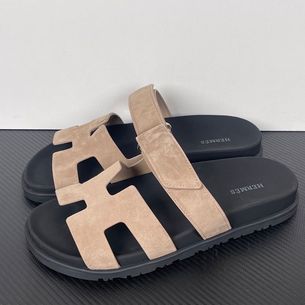 Angled top-down view of the Hermès Genius Sandals in taupe grey suede, showcasing the signature 'H' cutout front strap, the adjustable hook-and-loop instep strap, and the contoured black ergonomic rubber footbed with Hermès branding.