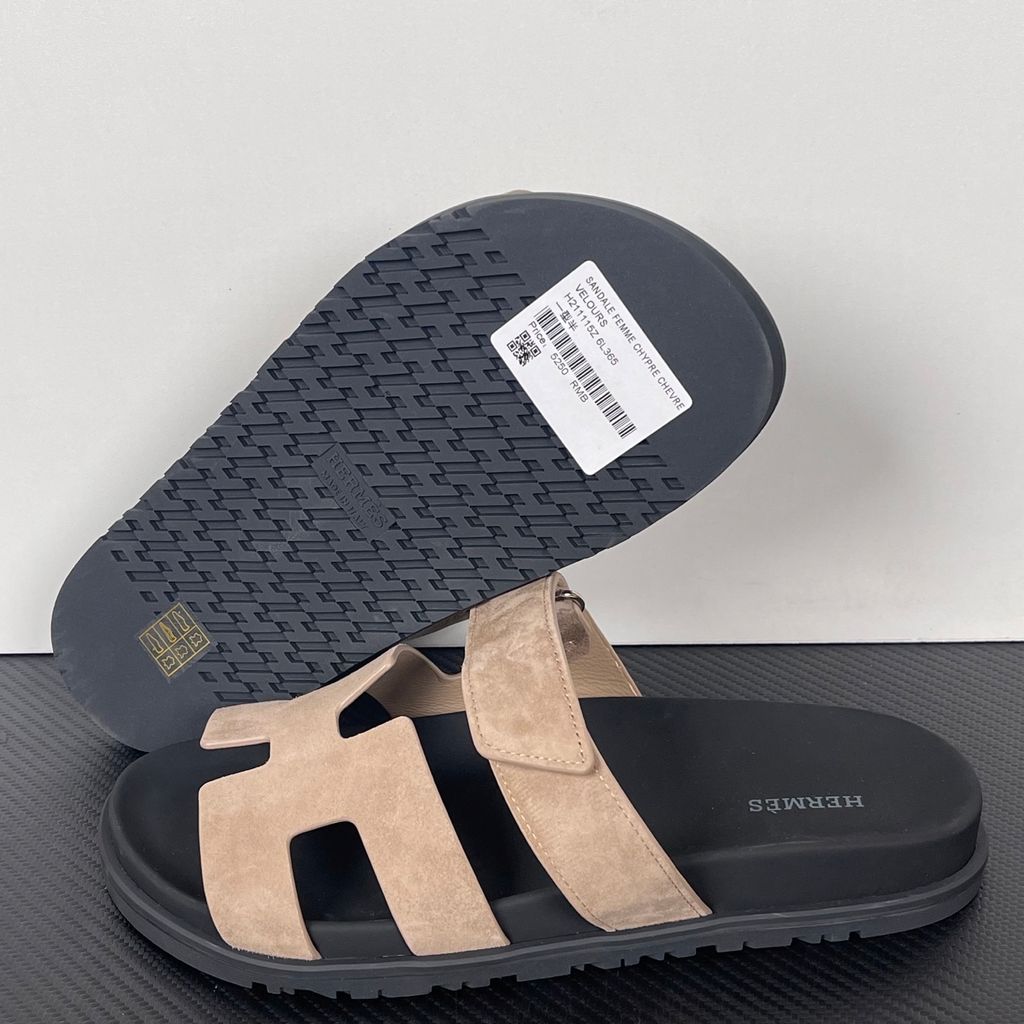 Angled top-down view of the Hermès Genius Sandals in taupe grey suede, showcasing the signature 'H' cutout front strap, the adjustable hook-and-loop instep strap, and the contoured black ergonomic rubber footbed with Hermès branding.