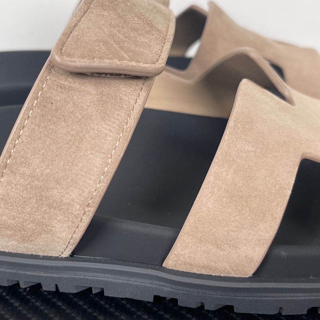 Angled top-down view of the Hermès Genius Sandals in taupe grey suede, showcasing the signature 'H' cutout front strap, the adjustable hook-and-loop instep strap, and the contoured black ergonomic rubber footbed with Hermès branding.